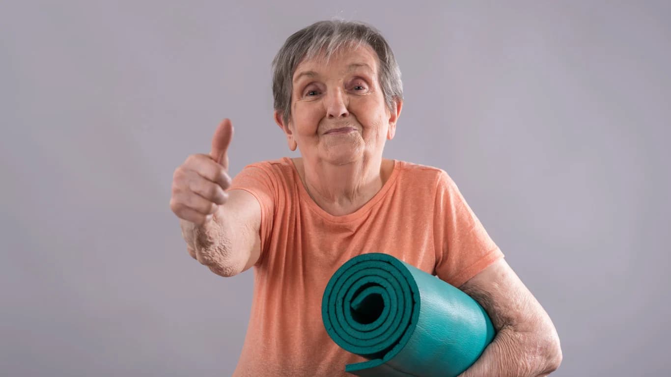 En äldre dam, som gör tumme upp med ena handen, pch bär på en hoprullad grön yogamatta under den andra armen